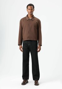 Bruine korte jas met een kraag, twee voorzakken en zwarte rechte jeans. Model staat tegen een neutrale achtergrond.