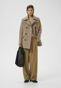 Cappotto doppio petto beige in lana con grandi bottoni neri, abbinato a pantaloni beige a vita alta e una borsa a tracolla nera.