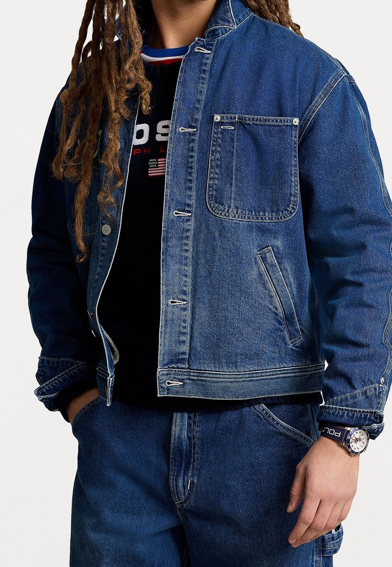 Persona che indossa una giacca di jeans blu e jeans, una maglietta nera con testo visibile e un orologio con cinturino scuro.