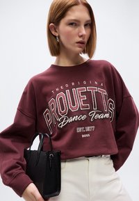 Μπορντό φούτερ με το κείμενο "PROUETTS Dance Team", φαρδιά μανίκια και στρογγυλή λαιμόκοψη. Μαύρη τσάντα με υφή κρατιέται στο αριστερό χέρι.