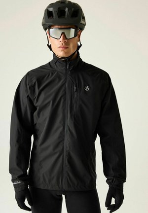 Mann trägt schwarzen Fahrradhelm, verspiegelte Sonnenbrille, schwarze Jacke und Handschuhe, schaut nach vorne vor einfachem Hintergrund.