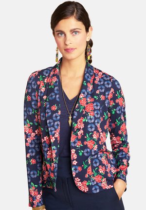 Woman with dark hair wears colorful floral blazer, navy shirt, pendant necklace, and long multicolored earrings, with hand in pocket.