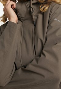 Veste légère marron à capuche avec cordon de serrage ajustable. Texture lisse, avec détails de coutures et logo sur la manche.