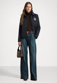 Giacca trapuntata blu navy con logo, dolcevita marrone, jeans a gamba larga blu e borsa marrone. Texture lisce e colori a contrasto.