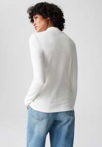 Weißes Langarmshirt mit weicher Textur und hohem Ausschnitt. Kombiniert mit hellblauen Jeans mit sichtbaren Gesäßtaschen.