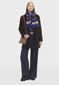 Brown wool coat, blue striped scarf, navy sweater, loose dark jeans, and brown bag. Model stands on a white background.