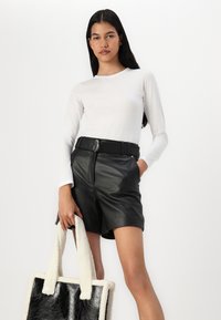 Un haut blanc à manches longues associé à des shorts en cuir noir taille haute. Tenant un sac fourre-tout texturé crème et noir.