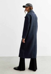 Cappotto in misto lana blu navy con colletto alto, maniche lunghe e taglio dritto, abbinato a pantaloni neri a campana e a un cappello grigio.