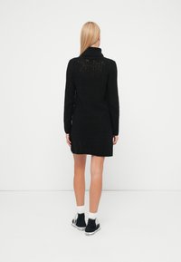 Juoda megzta suknelė su tekstūruota rombų rašto dekoracija, ilgomis rankovėmis ir aukštu apykakliu. Turi šiek tiek platėjančią siluetą.