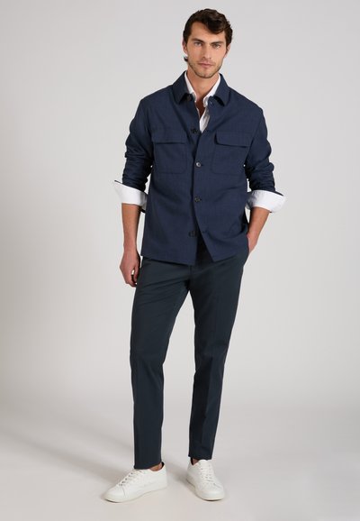 Man wearing navy buttoned jacket over white shirt with rolled sleeves, dark pants, and white sneakers standing against plain background.