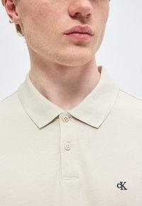 Jeune homme portant un polo beige à deux boutons avec un petit logo noir "cK" sur la poitrine, sur fond uni.