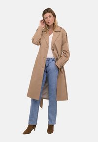 Cappotto trench beige con grandi bottoni, tasche laterali e epaulettes sulle spalle, indossato sopra una maglietta bianca e jeans blu a gamba dritta. Stivaletti marroni con tacco.