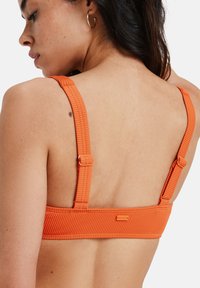 Top de bikini laranja com textura canelada, alças ajustáveis e acabamento em metal, apresentando um detalhe do logo na banda. Textura suave e design estruturado.