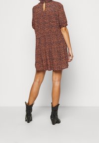Kortärmad klänning i knä längd i mörk orange med ett blommönster. Har en nyckelhålsknäppning och texturerad tyg. Bärs med svarta klackstövlar.