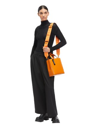 Woman in black turtleneck and wide pants holding orange handbag with wide branded strap on white background.