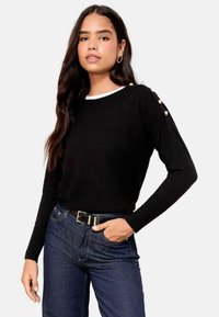 Maglione nero lavorato a maglia con polsini a coste, dotato di bottoni sulle spalle. Abbinato a jeans blu scuri e una cintura nera con una fibbia lucida.