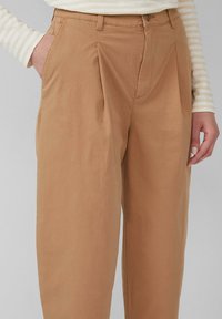Pantaloni plissettati color marrone chiaro con una consistenza morbida, dotati di tasche laterali e chiusura a bottone. Abbinati a un top a maniche lunghe a righe color crema e beige.