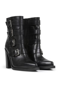 Botas negras hasta el tobillo hechas de cuero que cuentan con cuatro correas ajustables con hebillas plateadas, una puntera cuadrada y un tacón alto y grueso.