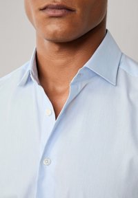 Kratki okvir donjeg dijela muškog lica i vrata na kojem je svijetloplava košulja s otvorenim gornjim gumbom.