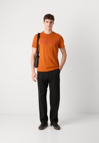 Oransje bomull T-skjorte med "BOSS" logo, kombinert med svarte bukser og svarte sko, holder en skulderveske. Modellen står foran en ensfarget bakgrunn.