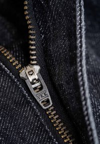 Zipper detail featuring dark metal pull tab labeled "YKK", surrounded by woven black fabric with visible texture and stitching accents.