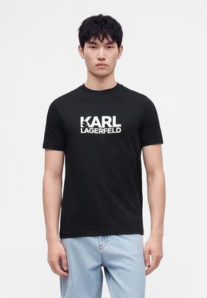 Giovane uomo che indossa una t-shirt nera Karl Lagerfeld e jeans azzurri, in piedi davanti a uno sfondo bianco semplice.