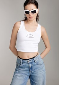 Top corto blanco con texto impreso, combinado con jeans de corte alto de color azul claro. La modelo lleva gafas de sol blancas y un sujetador negro debajo.