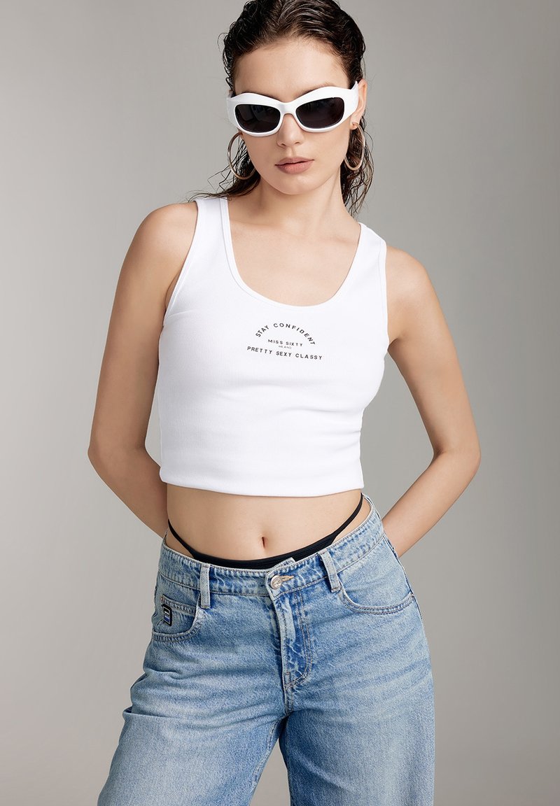 Weißes, kürzes Tanktop mit gedrucktem Text, kombiniert mit hellblauen, hochgeschnittenen Jeans. Das Model trägt weiße Sonnenbrillen und ein schwarzes Bralette darunter.