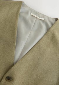 Oliivroheline vest tekstuurse kangaga, sileda halli voodriga ja ees pruun nööp. Märkimisväärne V-kujuline krae disain.