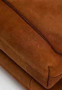 Bruine suède tas met een zachte textuur, gladde randen en een geperst merkteken. Compact rechthoekig formaat met zichtbare stiksels.