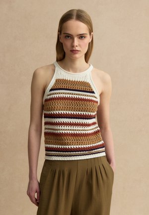 Joven mujer con cabello rubio liso, que lleva un top de punto sin mangas a rayas en tonos neutros y pantalones plisados oliva, de pie frente a un fondo beige.