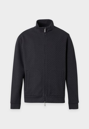 Mann steht, trägt eine schwarze Jacke mit Reißverschluss, weite schwarze Hose und schwarze Schuhe vor einem einfarbigen weißen Hintergrund.