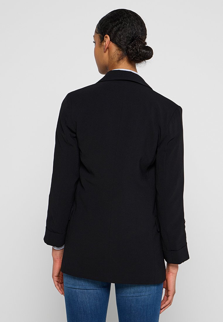 Blazer noir avec un revers cranté, des manches longues et des poignets retroussés, présentant une texture lisse et une coupe ajustée à l'arrière.