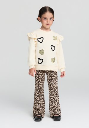Felpa chiara con maniche a volant, stampe a cuori neri e dettagli in paillettes dorate, abbinata a pantaloni a zampa d'elefante in stampa leopardo e scarpe nere.