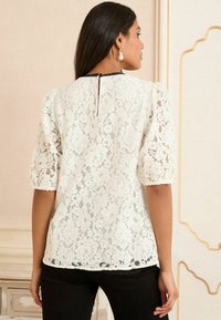 Top in pizzo color avorio con maniche a sbuffo, dotato di una apertura a chiave sul retro e rifiniture nere sul collo. Motivo in pizzo floreale con orlo scalloped.