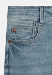 Jeans de mezclilla azul claro que presentan un pequeño bolsillo frontal, remaches de cobre y detalles de costura visibles. Textura suave con un acabado desgastado.