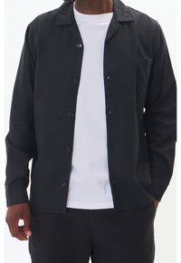 Czarna koszula zapinana na guziki z kołnierzykiem, długim rękawem i kieszenią na piersi, wykonana z teksturowanej tkaniny, noszona na białej koszulce.