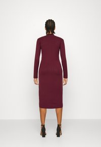 Person med flätat hår som bär en långärmad, ribbad, vinröd midiklänning och svarta högklackade sandaler, stående mot en vit bakgrund.