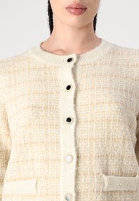 Cardigan beige texturé avec un col rond, décoré de grands boutons noirs, de deux poches avant et d'un tissu doux et duveteux.