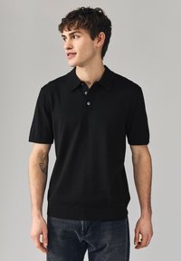 Polo noir à manches courtes avec col classique, doté d'une patte à trois boutons et d'un ourlet côtelé. Porté avec un jean en denim foncé.