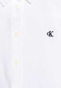Witte katoenen polo-shirt met een klassieke kraag, voorzien van vier knopen en een klein zwart geborduurd "cK"-logo op de linkerborst.