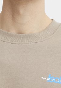 Sweatshirt en coton beige avec un col côtelé, arborant un motif "TOKYO" bleu et blanc. Le tissu a une texture lisse et une coupe décontractée.