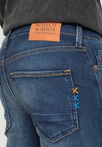 Tmavě modré džínové rifle s oranžovou koženou nášivkou s nápisem "SCOTCH & SODA AMSTERDAM". U kapes jsou barevné prošité detaily.