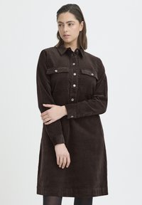 Mlada žena smeđe kose dužine do ramena nosi tamnosmeđu kordiranu haljinu na kopčanje s prednjim džepovima, stoji prekriženih ruku.