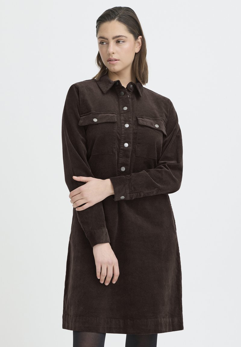 Mlada žena smeđe kose dužine do ramena nosi tamnosmeđu kordiranu haljinu na kopčanje s prednjim džepovima, stoji prekriženih ruku.