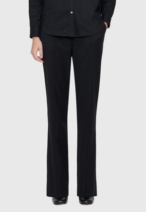 Person wearing black wide-leg trousers, black button-up shirt, and black shoes, standing with one hand in pocket against light background.