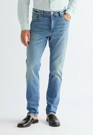 Mężczyzna w dopasowanych jasnoniebieskich dżinsach, jasnoniebieskiej koszuli z długim rękawem i czarnych wsuwanych butach, stoi z jedną ręką w kieszeni.