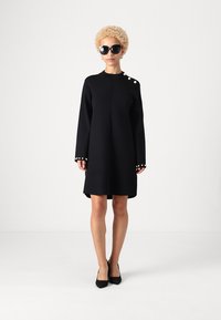 Čierne pletené šaty s dlhými rukávmi, s dekoračnými gombíkmi na jednom ramene a na manžetách. Kombinované s čiernymi podpätkami a slnečnými okuliarmi.