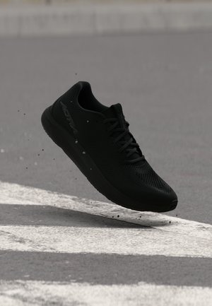Black running shoe with laces mid-air above a paved road with white crosswalk markings.