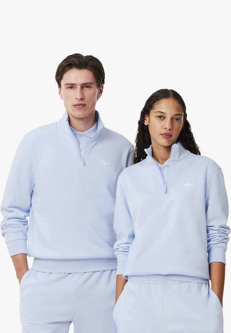 Felpe a pullover blu chiaro con colletto a zip, logo ricamato sul petto e polsini a costine. Entrambi gli individui indossano pantaloni abbinati.